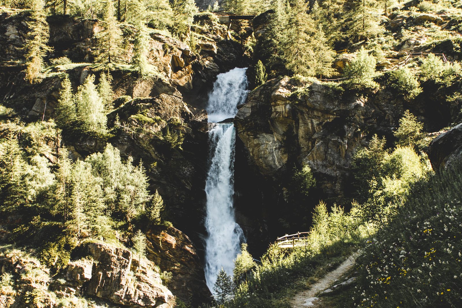 Sentiero alpino tra pini con cascata del Rutor sullo sfondo in Valle d'Aosta Sentiero alpino tra pini con cascata del Rutor sullo sfondo in Valle d'Aosta